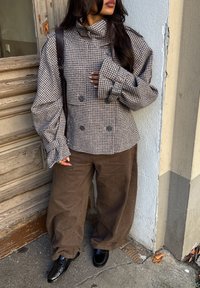 Brun houndstooth-mønstret oversized frakk med høy krage og dobbelbrytere, kombinert med brede brune kordfløyelsbukser og svarte sko.