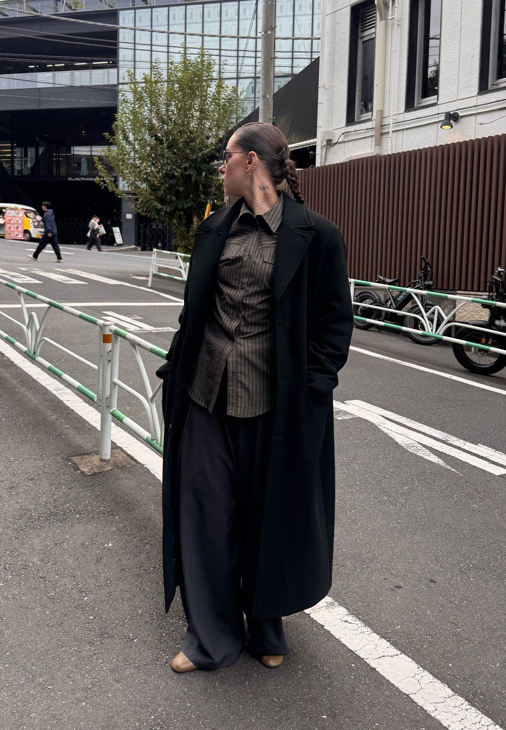 Long manteau noir, chemise à rayures boutonnée, pantalons larges foncés et chaussures beiges. Porte des lunettes de soleil élégantes et une coiffure tressée. Cadre urbain.