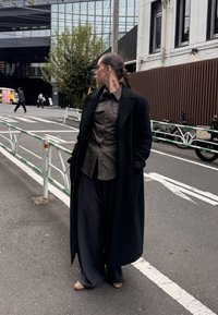 Long manteau noir, chemise à rayures boutonnée, pantalons larges foncés et chaussures beiges. Porte des lunettes de soleil élégantes et une coiffure tressée. Cadre urbain.