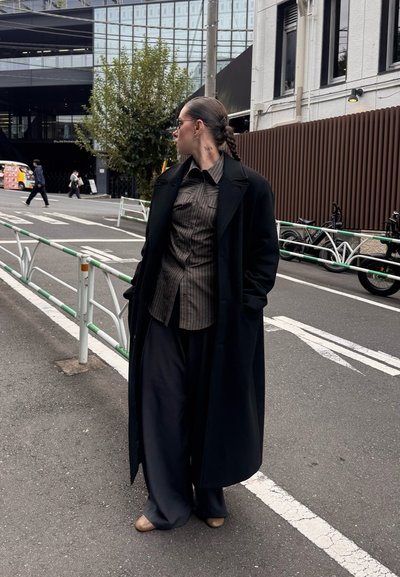 Abrigo largo negro, camisa de botones a rayas, pantalones anchos oscuros y zapatos beige. Lleva gafas de sol elegantes y un peinado trenzado. Ambiente urbano.