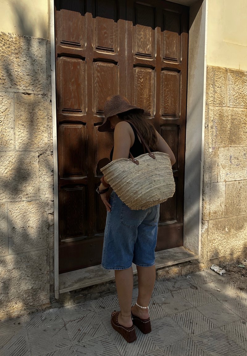 Υφασμένη τσάντα από ψάθα με καφέ δερμάτινες λαβές, σε υπερβολικό μέγεθος; φορεμένη με τζιν σορτς και καφέ πλατφόρμες μπροστά σε μια υφασμένη ξύλινη πόρτα.