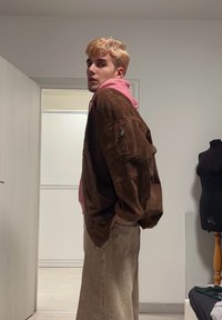 Veste oversized en daim marron avec une poche zippée, superposée sur un hoodie rose, associée à un pantalon baggy de couleur claire.