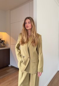 Blazer vert olive cintré avec deux boutons à l'avant et col à cran, associé à un pantalon large assorti, présenté dans un intérieur minimaliste.