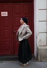 Blazer beige avec des épaules structurées sur une chemise blanche, associé à une longue jupe noire et des baskets marron, devant un fond de porte rouge.