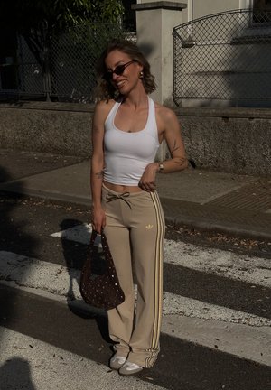 Mujer con top halter blanco, pantalones de chándal Adidas beige, zapatillas blancas, gafas de sol, sosteniendo un bolso marrón con tachuelas, de pie en un paso de peatones.