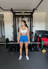 Barre d'haltères avec poids verts, sol en caoutchouc, équipement de fitness en arrière-plan, personne en haut blanc et short bleu, tenant la barre d'haltères.