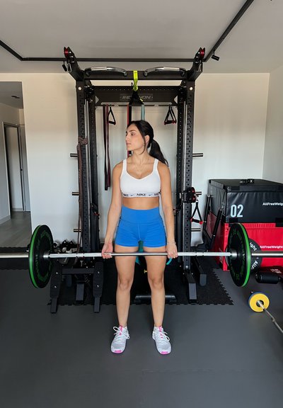 Barra con pesas verdes, suelo de goma, fondo con equipo de fitness, persona con camiseta blanca y pantalones cortos azules, sosteniendo la barra.