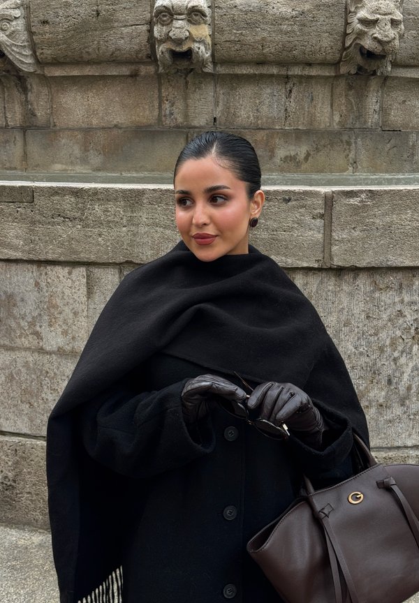 Une femme portant une cape noire et des gants en cuir tient des lunettes de soleil et un sac à main brun devant un mur de pierre sculptée.
