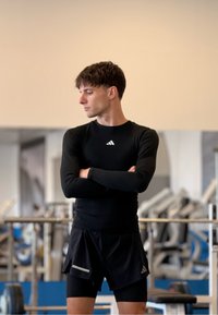 Fekete hosszú ujjú sportpulóver fehér logóval, fekete rövidnadrággal párosítva. A háttérben edzőtermi felszerelések láthatók.