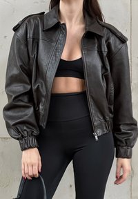 Mujer que lleva una chaqueta de cuero cortada negra sobre un sujetador deportivo negro y leggings negros de cintura alta, sosteniendo un bolso negro.