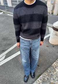 Crni i sivi prugasti pleteni džemper s okruglim izrezom, u kombinaciji s poderanim plavim trapericama i crnim cipelama. Mekani, teksturirani materijal.