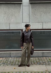 Braune, kurze Lederjacke mit Fronttaschen, kombiniert mit hellbraunen, weit geschnittenen karierten Hosen und einer strukturierten braunen Handtasche.