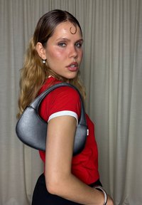 Ung kvinna med vågigt blont hår bär en röd skjorta, svart väska på axeln, silvriga örhängen och armband, poserar mot en enkel gardinbakgrund.