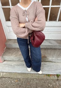 Cardigã rosa claro de malha com decote em V e detalhe de laço, combinado com jeans azul-escuro de perna larga e uma bolsa vermelha texturizada.