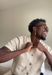 Rövid, göndör hajú, fülbevalós férfi nevet, miközben begombolja a krémszínű kötött rövid ujjú kardigánját, bent ülve egy sima fal előtt.