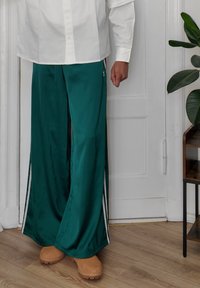 Pantaloni largi din satin verde, cu dungi laterale albe, completați de saboți bej. Textură netedă, croi lejer și design până la glezne.