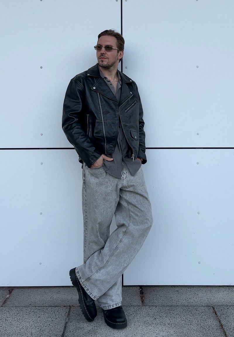 Nero giubbotto di pelle da motociclista, camicia grigia, jeans a gamba larga grigio chiaro e scarpe chunky nere. Il soggetto è appoggiato a un muro bianco.