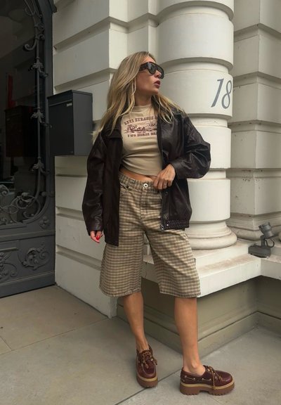 Chaqueta de cuero marrón, top corto beige con texto, pantalones cortos de cuadro y zapatos de plataforma burdeos con suelas marrones. Entorno al aire libre junto a un edificio.