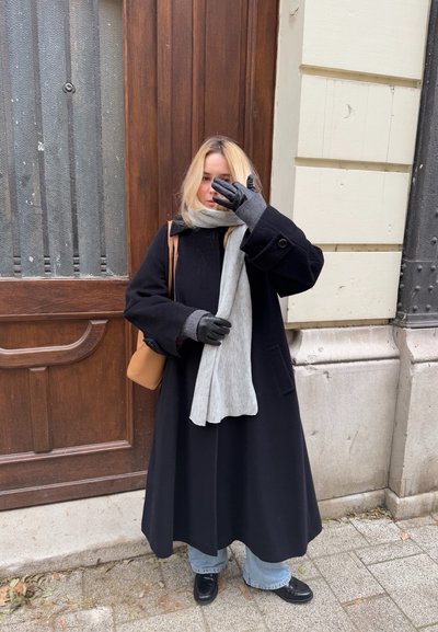 Abrigo largo negro, bufanda gris, guantes negros y zapatos oscuros, combinados con jeans de pierna ancha azul claro; de pie frente a una puerta de madera.