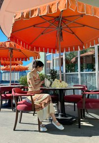 Φορεμα με σχέδιο σε κρέμα, κόκκινο και καφέ σε ζιγκ-ζαγκ; λευκά επίπεδα παπούτσια; περιτριγυρισμένο από πορτοκαλί ομπρέλες και κόκκινες καρέκλες σε ένα τραπέζι καφετέριας.