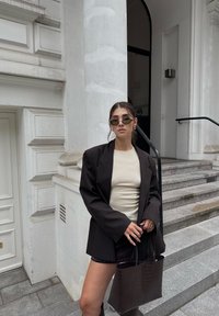 Blazer oversized con rayas marrones, top ajustado beige, pantalones cortos a juego con tachuelas, bolso tote negro texturizado, gafas de sol estrechas. Fondo de escalones de piedra.