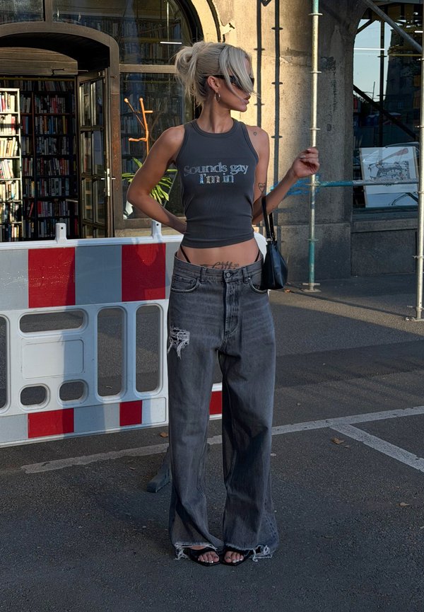 Persona con cabello corto y rubio que lleva una camiseta sin mangas y vaqueros anchos rasgados, de pie en la calle cerca de una barricada y un edificio con grandes ventanas.