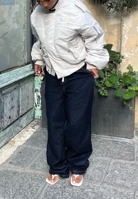 Light grey puffer jacket, dark wide-leg jeans, and white flip-flops. Textured materials and relaxed fit. Standing near plants and a weathered wall.