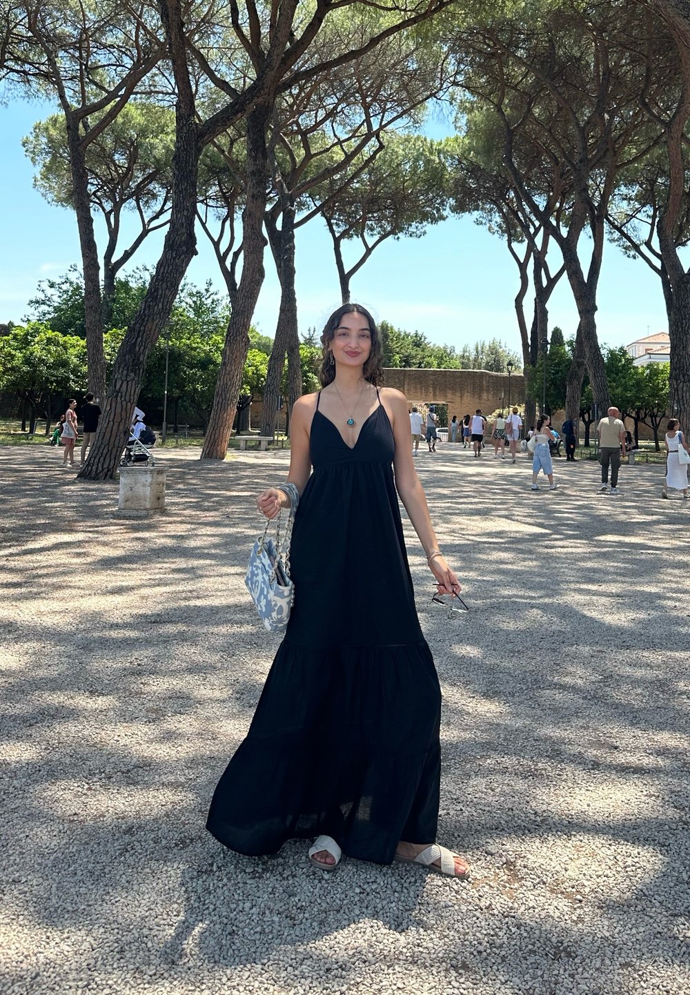 Vestido maxi preto, sem mangas, com saia em camadas, combinado com sandálias brancas e uma bolsa estampada azul clara. Luz do sol filtrando através das árvores.