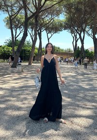 Vestido maxi preto, sem mangas, com saia em camadas, combinado com sandálias brancas e uma bolsa estampada azul clara. Luz do sol filtrando através das árvores.