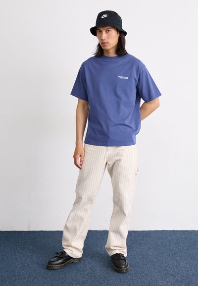 Camiseta de algodón azul, de corte relajado, con texto blanco. Pantalones de rayas beige claro con bolsillos laterales. Sombrero tipo bucket negro y zapatos negros con cordones.