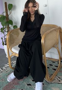Haut noir à manches longues avec des accents en dentelle au niveau du col, associé à un pantalon noir ample et des baskets blanches, assis sur une chaise en rotin.