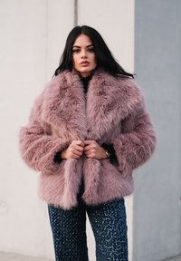 Palton din blană sintetică de culoare lavandă deschisă, cu guler larg și textură pufoasă; asortat cu pantaloni bleumarin cu paiete, creând un contrast între materiale.