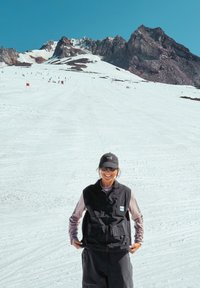 Vesta neagră fără mâneci peste o cămașă cu mânecă lungă roz, stând pe zăpadă cu un fundal montan și schiori în depărtare. Cer albastru strălucitor.