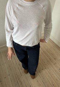 Personne portant une chemise blanche à manches longues et un jean foncé, debout sur un sol en bois clair, une main sur la hanche et l'autre détendue.