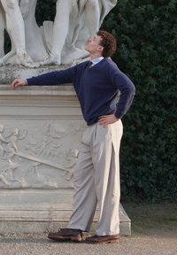 Giovane in un maglione blu navy e pantaloni grigi chiari, in piedi vicino a una statua di pietra bianca con figure scolpite, che mostrano dettagli intricati.