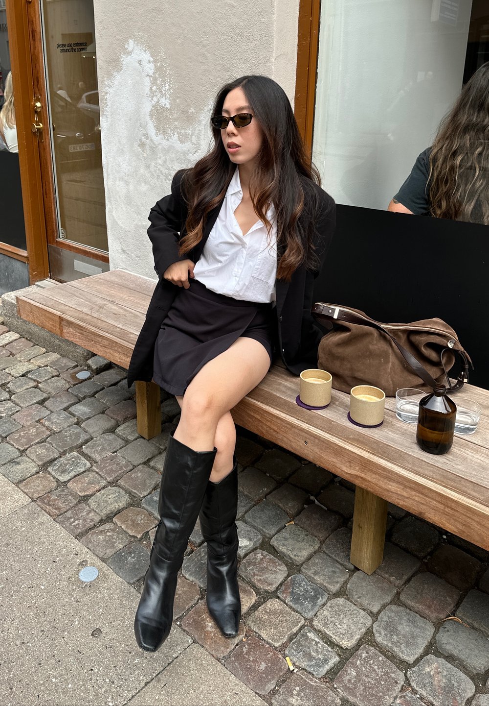 Schwarze Lederstiefel bis zum Knie, weißes Hemd, schwarze Blazer und Rock, sitzend auf einer Holzbank mit Kaffeetassen und einer braunen Tasche in der Nähe.
