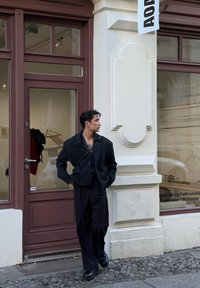 Μαύρο κοντό σακάκι με μεγάλο γιακά πάνω από μια μαύρη μπλούζα, συνδυασμένο με χαλαρούς μαύρους παντελόνες και χοντρές μαύρες μπότες, έξω από ένα μαγαζί.