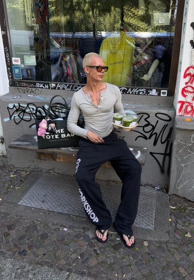 Bolso tote negro con texto blanco, camiseta de manga larga gris, pantalones negros y chanclas. Tres bebidas verdes en una bandeja. Graffiti en el fondo.