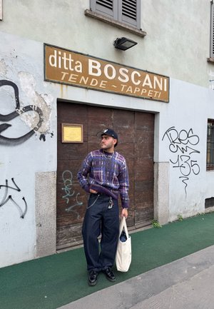 Pull en motif violet, pantalon large en denim sombre, chaussures noires et un sac en toile blanc. L'arrière-plan présente une porte en bois et des graffitis sur les murs.