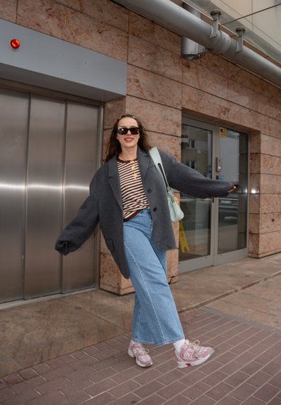 Mujer con abrigo gris de gran tamaño, camisa de rayas, vaqueros de pierna ancha y zapatillas rosas, de pie con los brazos extendidos cerca del ascensor y las puertas de cristal.