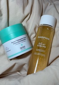 Feuchtigkeitscreme mit einem türkisfarbenen Deckel, beschriftet mit "Protini." Goldene Behandlungessenz in einer durchsichtigen Flasche, beschriftet mit "Éclat Sublime."