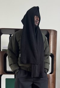 Homme portant un bonnet noir et une longue écharpe noire sur une veste matelassée verte, debout les mains dans les poches devant une étagère en bois.