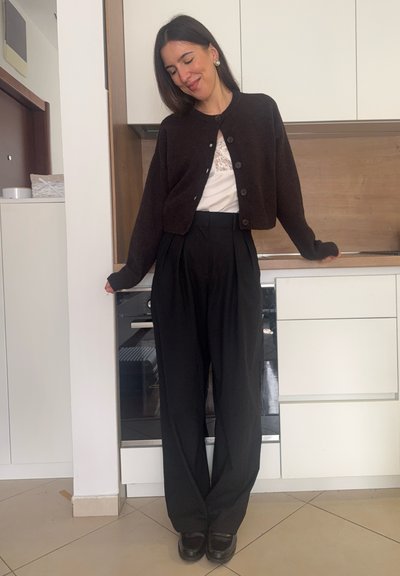 Mujer de pie en una cocina, vistiendo un cárdigan negro, blusa blanca de encaje, pantalones negros de talle alto y zapatos negros, con los ojos cerrados y sonriendo.