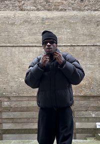 Schwarze Daunenjacke mit hohem Kragen, getragen über einem schwarzen Outfit. Accessoires umfassen dunkle Sonnenbrillen und mehrere Ringe. Texturierten Hintergrund.