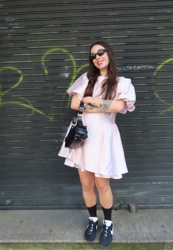 Donna con un vestito rosa pallido, sneaker nere, occhiali da sole e una borsa nera a tracolla, in piedi con le braccia incrociate davanti a una porta metallica coperta di graffiti.