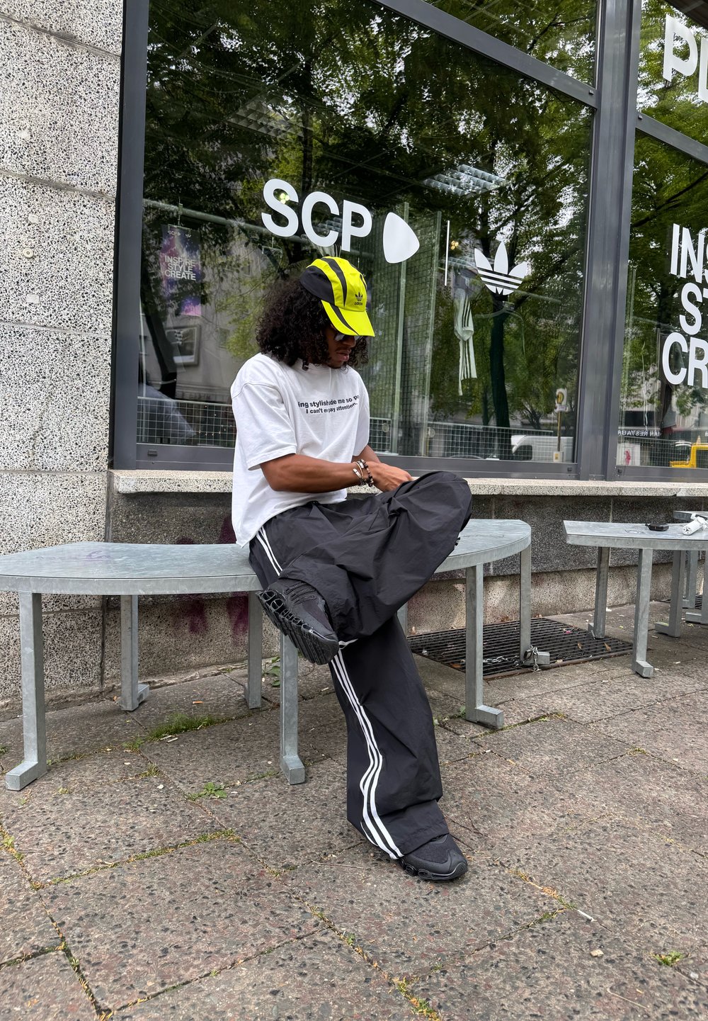 Personne portant une casquette jaune et noire, un t-shirt blanc à motif graphique et un pantalon noir avec des rayures blanches, assise sur un banc devant un magasin SCP.