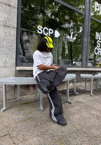Personne portant une casquette jaune et noire, un t-shirt blanc à motif graphique et un pantalon noir avec des rayures blanches, assise sur un banc devant un magasin SCP.
