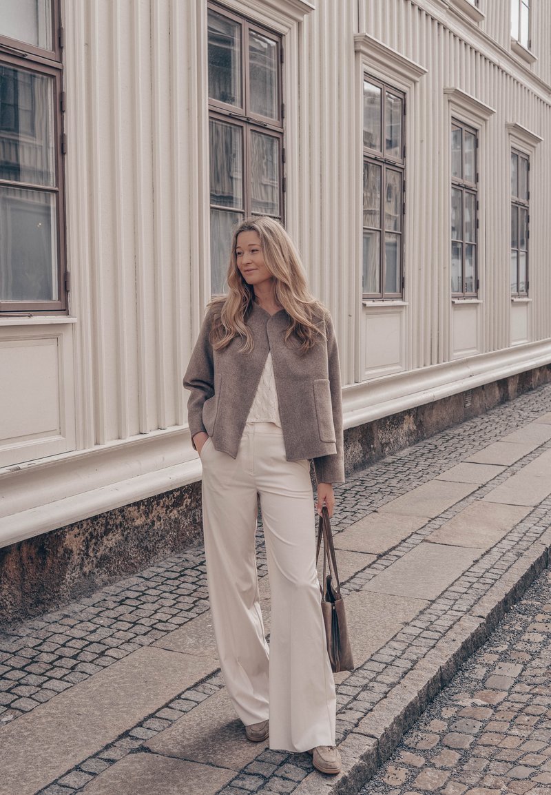 Femme aux longs cheveux portant un pantalon beige à jambes larges et une veste marron, debout dans une rue pavée à côté d'un bâtiment classique.