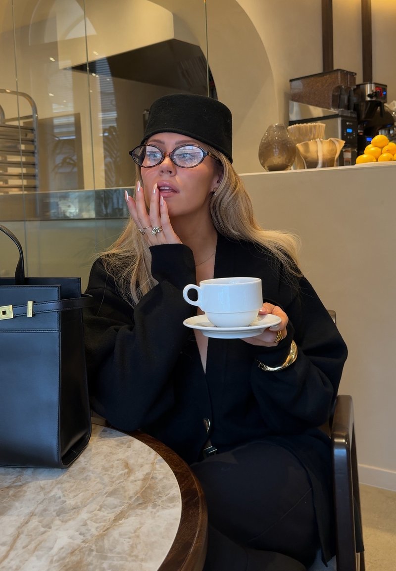 Femme portant un chapeau noir et des lunettes, assise à une table en marbre dans un café, tenant une tasse blanche et une soucoupe près du visage, avec un sac à main noir à côté d'elle.