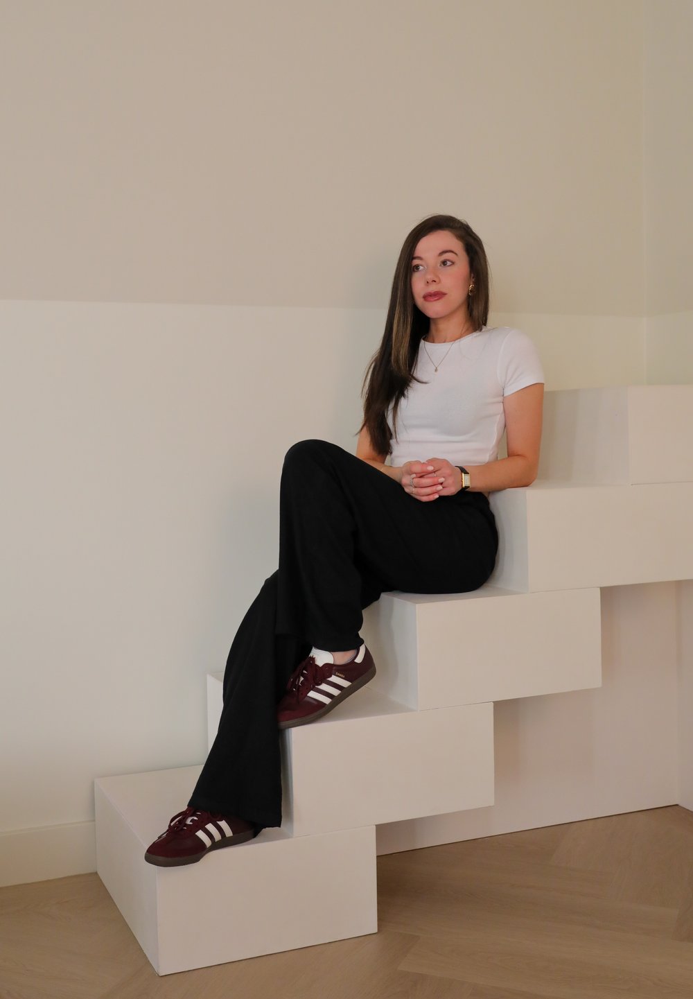 Hvit kortermet skjorte, svarte vide bukser og maroon sneakers med hvite striper. Sitter på hvite geometriske trinn. Nøytral bakgrunn.